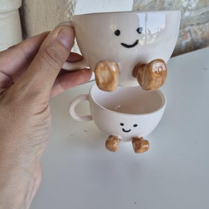 Könnte beinhalten: Zwei gestapelte, cremefarbene Keramik-Kaffeetassen mit lächelnden Gesichtern und braunen Füßen. Die obere Tasse ist etwas größer und ruht auf der unteren Tasse. Die Tassen haben Henkel.