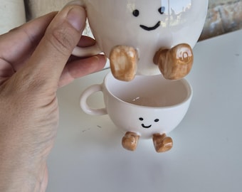 Puede incluir: Dos tazas de café apiladas de cerámica blanquecina con caras sonrientes y pies marrones. La taza superior es ligeramente más grande y descansa sobre la taza inferior. Las tazas tienen asas.