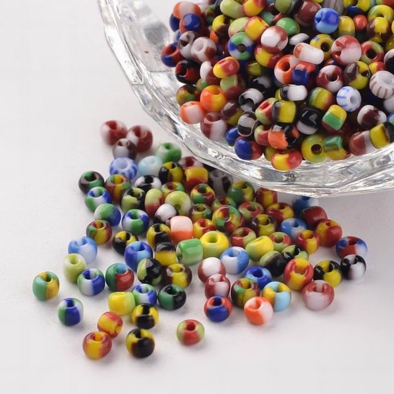 May include: A bowl filled with colorful glass seed beads. The beads are a mix of red, yellow, blue, green, white, and black. The beads are scattered on a white surface.