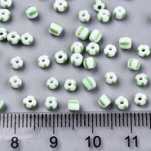 May include: A collection of white and green striped glass beads. The beads are approximately 6mm in diameter and are scattered on a gray surface. A ruler is visible in the bottom of the image.