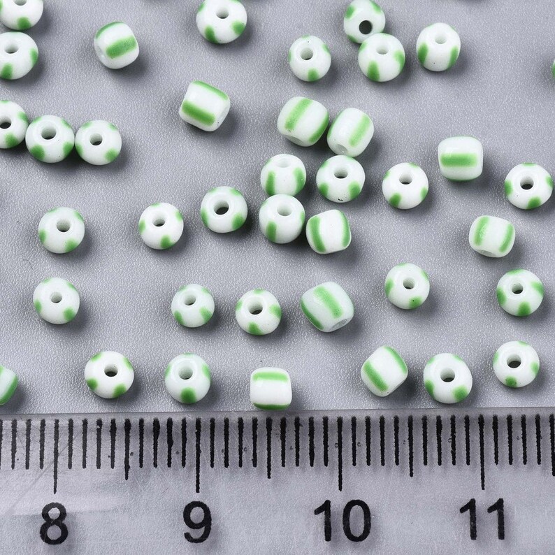 May include: A collection of white and green striped glass beads. The beads are approximately 6mm in diameter and are scattered on a gray surface. A ruler is visible in the bottom of the image.