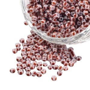 May include: A bowl of white and brown striped glass beads. The beads are small and round, and they are scattered on a white surface.