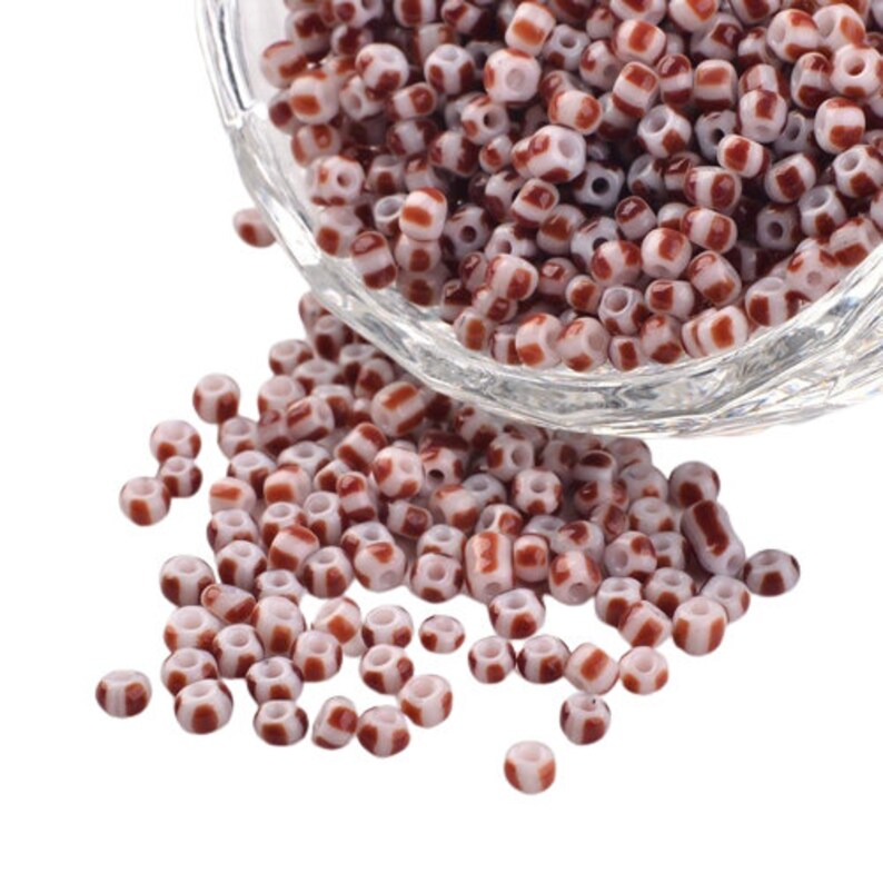 May include: A bowl of white and brown striped glass beads. The beads are small and round, and they are scattered on a white surface.