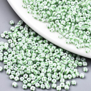 May include: A close-up of a large quantity of white and green glass seed beads. The beads are small and round, with a white base and green stripes.