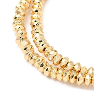 May include: Two strands of gold-colored faceted beads arranged in a curved formation. Each individual bead features a reflective, multi-faceted surface that catches the light, creating a bright, metallic appearance across the entire length.