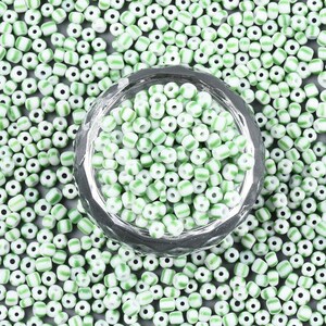 May include: A close-up of a glass bowl filled with white and green striped glass beads. The beads are scattered around the bowl on a gray surface.
