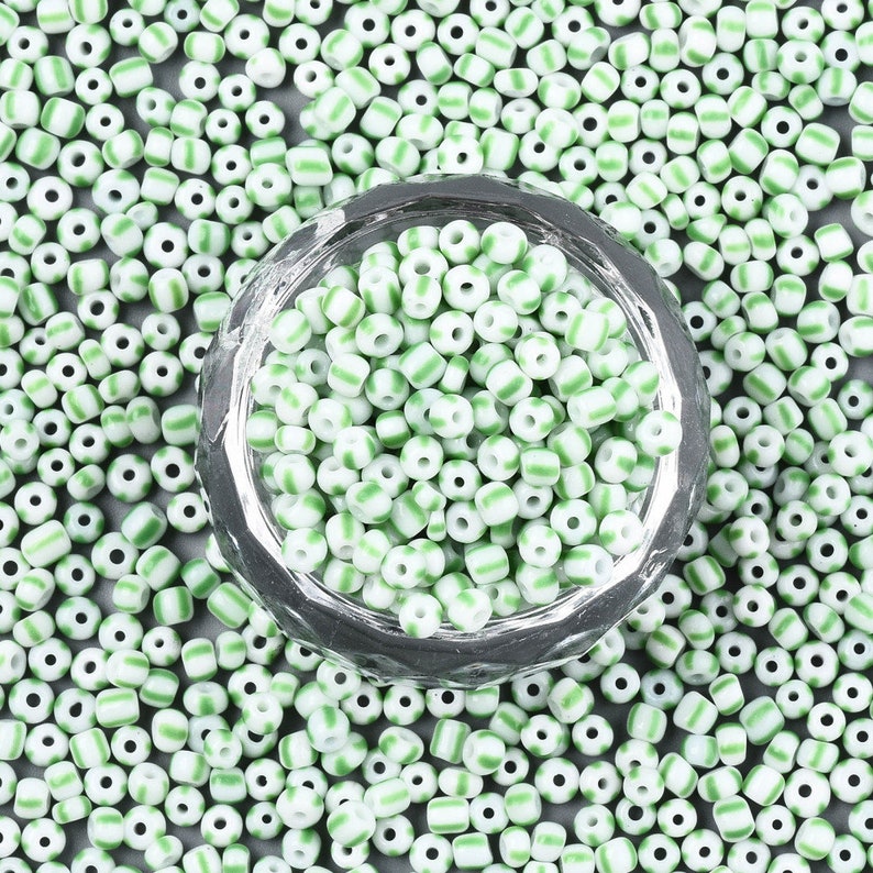 May include: A close-up of a glass bowl filled with white and green striped glass beads. The beads are scattered around the bowl on a gray surface.