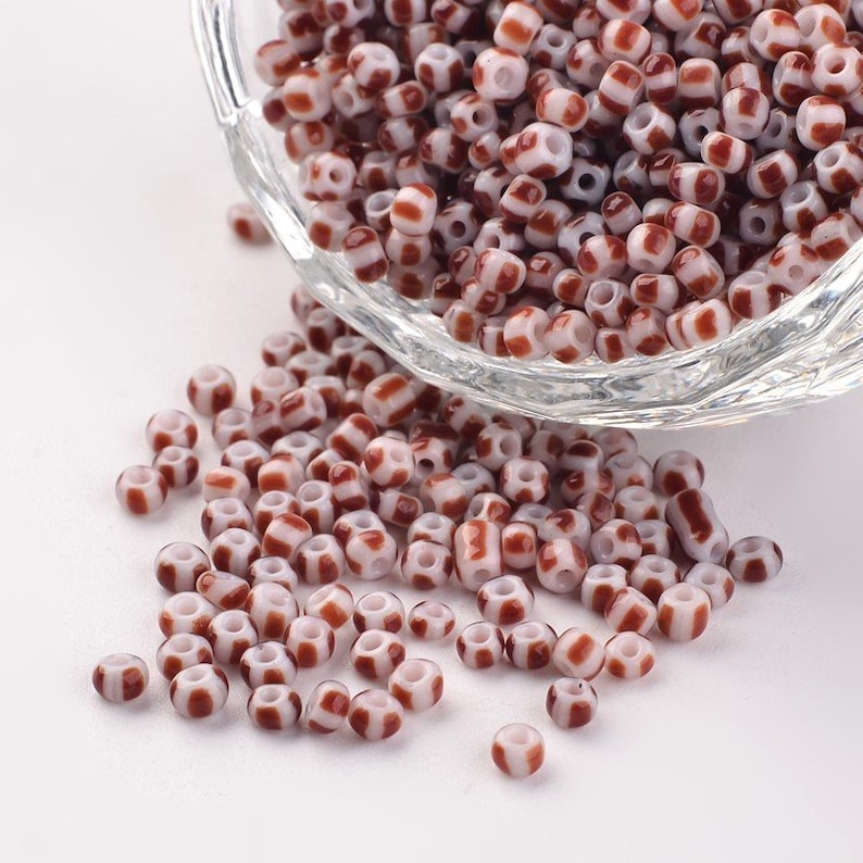 May include: A bowl of white and brown striped glass seed beads. The beads are small and round, and they are scattered on a white surface.