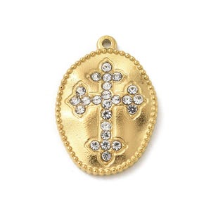 May include: An oval gold-toned metal pendant featuring a central cross motif embellished with clear, sparkling rhinestones. The pendant has a beaded border and a small loop at the top for attachment, showcasing a textured, matte finish.