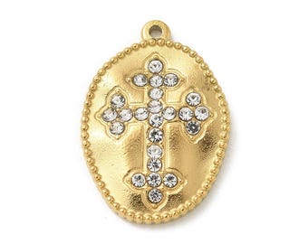 May include: An oval gold-toned metal pendant featuring a central cross motif embellished with clear, sparkling rhinestones. The pendant has a beaded border and a small loop at the top for attachment, showcasing a textured, matte finish.
