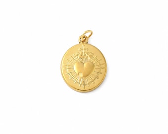 Puede incluir: Un colgante redondo de oro con un diseño en relieve de un corazón central coronado por una cruz, rodeado de líneas radiantes. El colgante incluye una pequeña anilla circular en la parte superior para sujetarlo a una cadena.