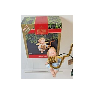 May include: A Hallmark Keepsake Sparkling Angel ornament featuring a light blue robe and gold tinsel garland. The box displays the text Magic Blinking Lights, Keepsake Ornament, and Sparkling Angel. The angel holds a gold tinsel garland.