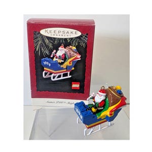 May include: A Christmas ornament featuring Santa Claus in a LEGO sleigh. The sleigh is blue, red, and yellow, with white runners. Santa is holding a candy cane and a sack of toys. The box reads "Keepsake Ornament 1999".