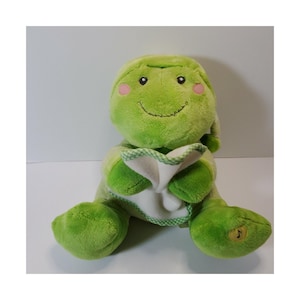 May include: A plush green frog toy with a smiling face, pink cheeks, and a white bib with a green and white checkered trim. The frog has large feet and a musical note symbol on one foot.