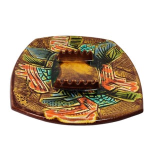 May include: A vintage ceramic ashtray with a unique, colorful design. The ashtray features a brown base with a geometric pattern in orange, green, and blue. A central, rectangular section holds the cigarette rests. The overall shape is a rounded square.