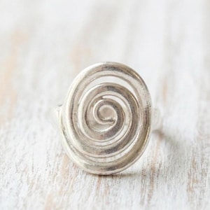 May include: A silver ring featuring a spiral design. The ring has an oval shape with a raised, openwork spiral in the center. The ring is set against a light-colored, textured background.