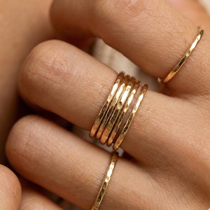Pode incluir: Close-up de vários anéis dourados com textura martelada. Os anéis são empilhados em um dedo, com alguns outros visíveis. Os anéis têm um design simples e elegante.