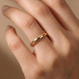 Könnte beinhalten: Ein goldfarbener Ring mit einem glatten, abgerundeten Band. Der Ring wird an einem Finger getragen, reflektiert Licht und zeigt seine polierte Oberfläche. Das Schmuckstück ist schlicht und elegant.