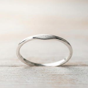 Può includere: Un anello d'argento con una parte superiore strutturata e leggermente appiattita. La fascia è liscia e arrotondata, con una curva sottile. L'anello è posto su uno sfondo chiaro e neutro, che ne sottolinea l'elegante semplicità. L'anello è probabilmente in argento sterling.