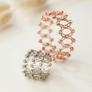 May include: Two expandable rings, one silver and one rose gold, each set with small, clear gemstones. The rings feature an intricate, openwork design. They are displayed on a white surface, highlighting their delicate and detailed construction.