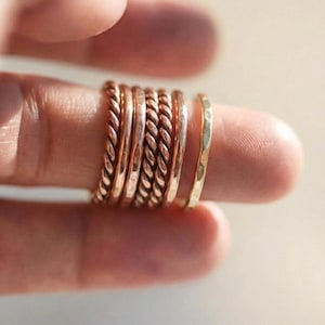 Könnte beinhalten: Eine Sammlung gold- und kupferfarbener Ringe. Mehrere Ringe haben ein gedrehtes Seil-Design, während andere glatte Bänder sind. Ein Ring hat eine gehämmerten Textur. Die Ringe sind auf einem Finger gestapelt und zeigen die verschiedenen Stile.