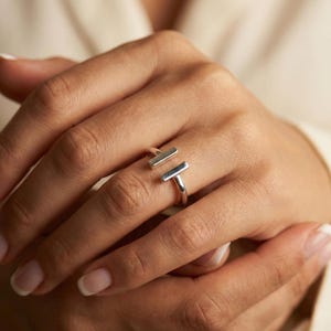 Op de afbeelding: Een zilveren ring met een modern design. De ring heeft twee parallelle rechthoekige staven die door een band zijn verbonden. De ring zit om een vinger, met een zachte, neutrale achtergrond. Een eenvoudig, elegant sieraad.
