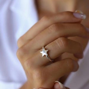 Puede incluir: Un anillo plateado con un diseño de estrella de cinco puntas. El anillo se lleva en un dedo, mostrando la forma de la estrella y la banda del anillo. Una pieza de joyería sencilla y elegante.