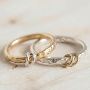 Può includere: Due anelli intrecciati con una texture martellata. Un anello è color oro e presenta due piccoli anelli color argento infilati attorno. L'altro anello è color argento, marchiato 925, e presenta due piccoli anelli color oro infilati attorno.