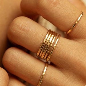 May include: Close-up of several gold-coloured rings with a hammered texture. The rings are stacked on a finger, with additional rings on adjacent fingers. The rings have a simple, elegant design.