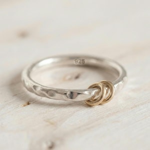 Puede incluir: Un anillo de plata con textura martillada y dos pequeños anillos dorados adjuntos. El anillo está marcado con "925". El anillo está sobre una superficie de madera clara.