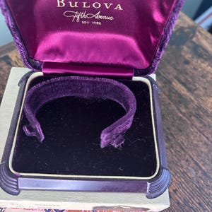 May include: A vintage Bulova Fifth Avenue New York jewelry box. The box is lined with purple velvet and features a matching velvet insert. The box is open, revealing the interior, and sits on a wooden surface.
