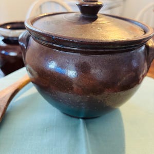 May include: A brown ceramic pot with a lid, featuring a rounded shape and two side handles. The pot has a glossy finish and a dark brown color. A second pot is partially visible in the background. The pot is sitting on a light blue surface.