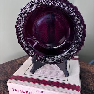 May include: A deep ruby red glass bowl with a scalloped edge and a textured diamond pattern on the rim. The bowl sits on a black plastic stand atop a white cardboard box labelled The 1876 Cape Cod Collection Avon Soup/Cereal Bowl.