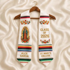 May include: White graduation stole with floral and religious imagery. Features the text "CLASS OF 2026", "ALEX GARCIA", and "ST. MARY'S COLLEGE". The stole has a colorful striped design at the bottom and is displayed on a wooden hanger.