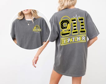 May include: Gray t-shirt with "911 DISPATCHER" design on the back, featuring a headset graphic and a heartbeat line. The front has a smaller logo with the text "DISPATCHER Team".