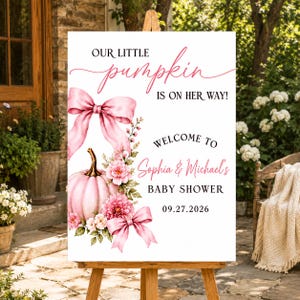 May include: A white rectangular baby shower welcome sign featuring a pink pumpkin, pink bows, and floral accents. The text reads: OUR LITTLE pumpkin IS ON HER WAY! WELCOME TO Sophia & Michael's BABY SHOWER 09.27.2026. The sign is on a wooden easel.