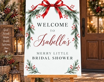 May include: A white rectangular sign on a wooden easel featuring a red ribbon bow at the top, green pine branches, and red berries. The text reads: WELCOME TO Isabella's MERRY LITTLE BRIDAL SHOWER in black and red serif and script fonts.