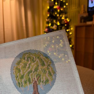 May include: Needlepoint canvas featuring a tree design. The tree has a brown trunk, green leaves, and is set against a light blue background within a circle. The canvas is white with a grid pattern.