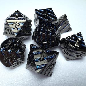 Puede incluir: Juego de siete dados de metal negro con detalles azules. Los dados son de múltiples caras, incluido un D20, y presentan números y diseños grabados. Para juegos de mesa.