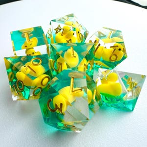 Pode incluir: Um conjunto de dados poliédricos com resina transparente verde-azulado. Cada dado apresenta um patinho de borracha amarelo no interior, com números dourados. Os dados estão dispostos em um grupo, mostrando seu design único e tema lúdico.