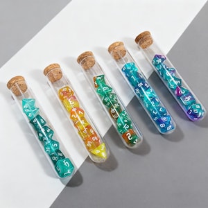 Peut inclure: Cinq éprouvettes en verre avec des bouchons en liège, chacune remplie de dés polyédriques colorés. Les dés sont de différentes couleurs, dont le vert, le jaune, le bleu et le violet. Les tubes sont disposés sur une surface blanche et grise.