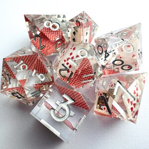 Può includere: Set di sette dadi in resina trasparente con immagini di carte da gioco e dettagli rossi. Ogni dado presenta numeri argentati e simboli di seme. Ideali per giochi da tavolo e di ruolo.