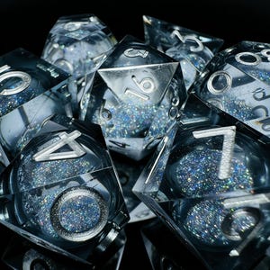 May include: A set of clear, multifaceted dice with silver numbering and embedded glitter. The dice are arranged on a black surface, reflecting light and creating a sparkling effect. Ideal for tabletop games.
