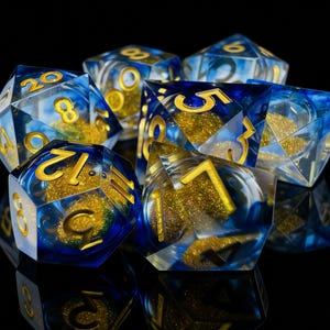 May include: A set of polyhedral dice with a blue and gold color scheme. The dice are transparent with gold glitter and blue swirls. The numbers are in gold. The dice are displayed on a black surface.