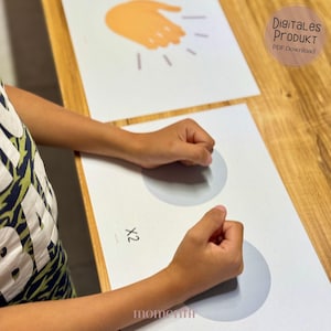 May include: A child's hands interacting with a printed educational game. The game features a hand illustration and two gray circles on white paper. The text "X2" is visible. The image also includes the text "Digitales Produkt PDF Download".
