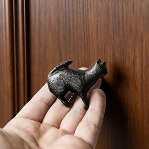 Pode incluir: Puxador de armário preto em forma de gato. O puxador é feito de um material texturizado e é segurado em uma mão. O gato está em uma pose de caminhada com a cauda para cima. O fundo é uma porta de armário de madeira.