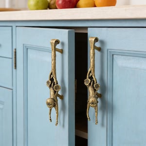 May include: Two bronze-colored cabinet handles shaped like monkeys hanging from a tree branch. The handles are mounted on light blue cabinet doors. The cabinet doors are open, revealing the interior.