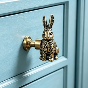 May include: A brass-colored cabinet knob featuring a detailed rabbit design. The knob is mounted on a light blue cabinet door. The rabbit is in a seated position with its ears upright.