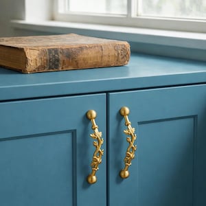 May include: A close-up of a blue cabinet with two gold-colored, leaf-design handles. An antique book with a worn, brown cover rests on top of the cabinet. The cabinet is positioned near a window.