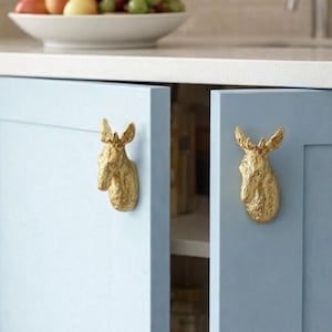 Peut inclure: Poignées de placard en forme de tête d'élan dorée, montées sur des portes de placard bleu clair. Les portes sont fermées, avec un plan de travail blanc et un bol de fruits en arrière-plan. Décoration rustique.